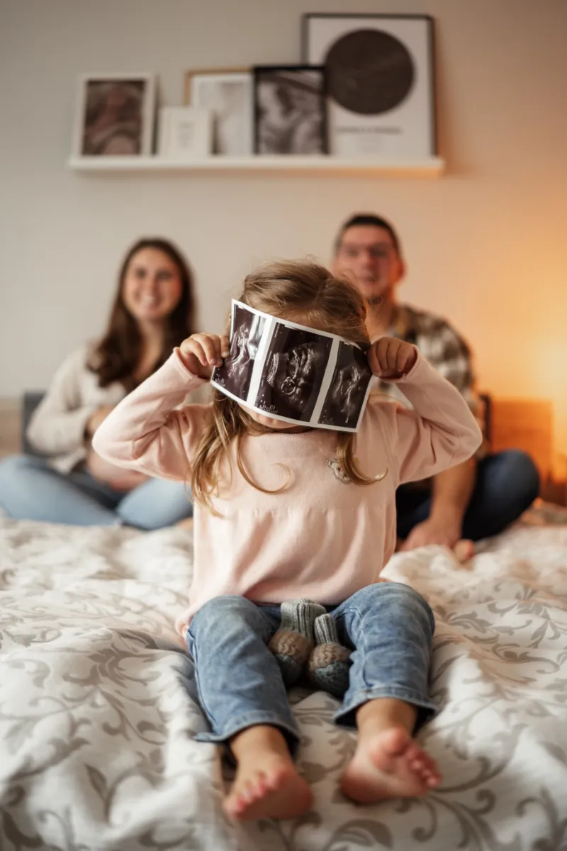 Große Schwester hält sich Ultraschallbilder vom Geschwisterchen vors Gesicht, Eltern lachend im Hintergrund